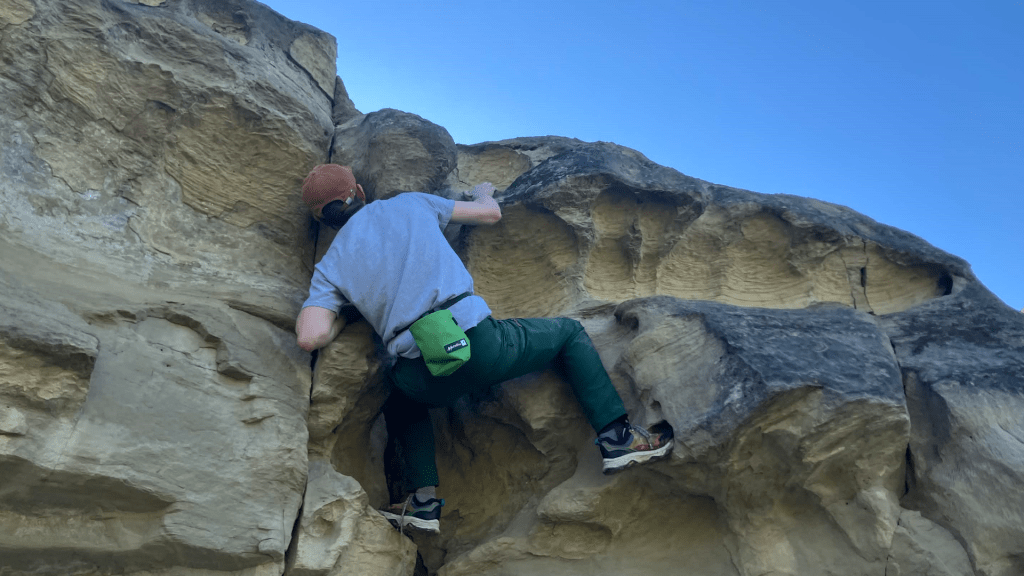 A person bouldering on a sandstone rock face, reaching for a handhold while wearing a chalk bag beneath a clear blue sky.